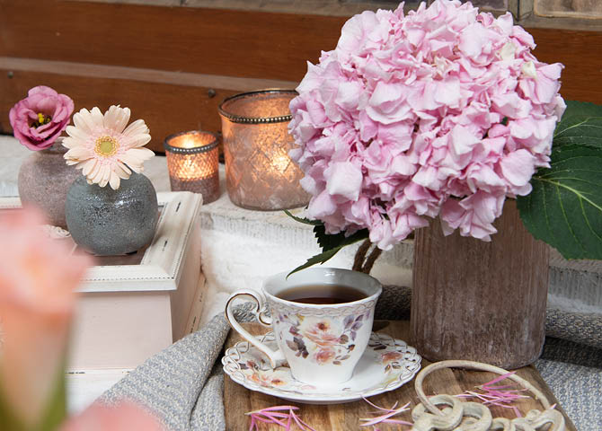 Un tagliere con una tazza e piattino, candele, vasi per fiori e una scatola del tè.