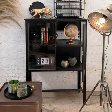 Ein Schrank mit einem Globus, Töpfen, einer goldenen Tigergussfigur, einem Buch und einem Tablett mit Kerzen und einer Lampe im industriellen Stil.