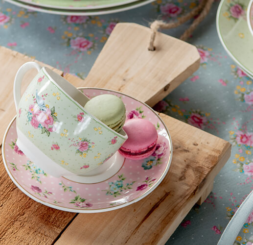 Zwei Macarons werden in einer Kaffeetasse und Untertasse mit einem romantischen Motiv präsentiert, wobei die Tasse schräg steht.
