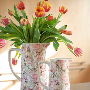 Tulpen in verschillende kleuren gepresenteerd in een romantische schenkkan met een rozenmotief in roze tinten. Aan de onderkant van de afbeelding bevindt zich een knop met het label 'Moederdag', die linkt naar de Moederdag pagina.