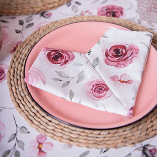 A pink plate resting on a beige seagrass placemat. On the plate are two napkins elegantly folded into a heart shape, featuring a pattern of pink roses.  At the bottom of the image is a button labeled 'Valentine's,' linking to the Valentine's page.