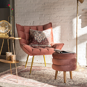 A homely image featuring a pink velvet armchair and a matching pink velvet pouf, both adorned with golden legs. Next to the armchair stands an elegant golden side table.