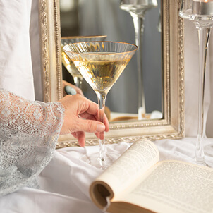 An image of a martini glass filled with prosecco, held by a woman. Only her hand is visible, covered in a lace sleeve. In the background, there is a white tablecloth with a golden mirror.