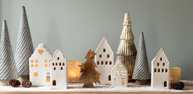 Una scena di decorazioni natalizie, tra cui un albero di Natale in legno, casette decorative in ceramica, piccoli alberi di Natale grigi, alberi di Natale dorati e portacandele gialli