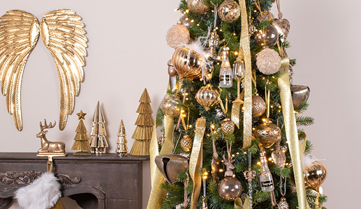 The golden Christmas trend with a Christmas tree adorned with gold ornaments and baubles, and a brown mantelpiece with a gold Christmas stocking hanger and three gold Christmas tree figures and golden wings on the wall
