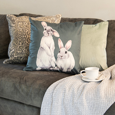 A gray fabric sofa with three decorative cushions in solid colors, featuring rabbits and oriental patterns. There's also a white throw with a cup and saucer on the sofa