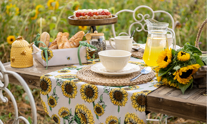 Ein gedeckter Tisch im Sonnenblumen-Thema mit einem Tischläufer, rustikalem Geschirr, runden Platzsets aus Weide, einem Brotkorb gefüllt mit Baguettes und einer Kanne