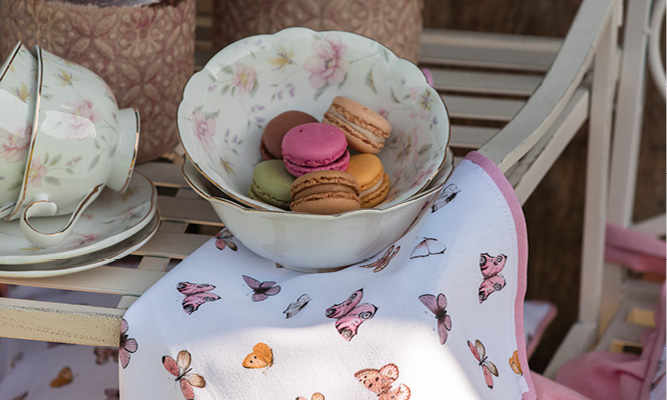 Eine antike Schale gefüllt mit bunten Macarons, darunter ein Baumwollserviette mit rosa Schmetterlingen