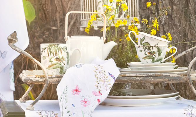 A colorful outdoor setting with a metal tray featuring botanical teacups and a mug, along with a white modern teapot