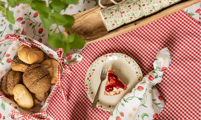A set table with a strawberry theme, including a bread basket filled with rolls, a napkin, a dessert plate with strawberry cake, and strawberry mugs