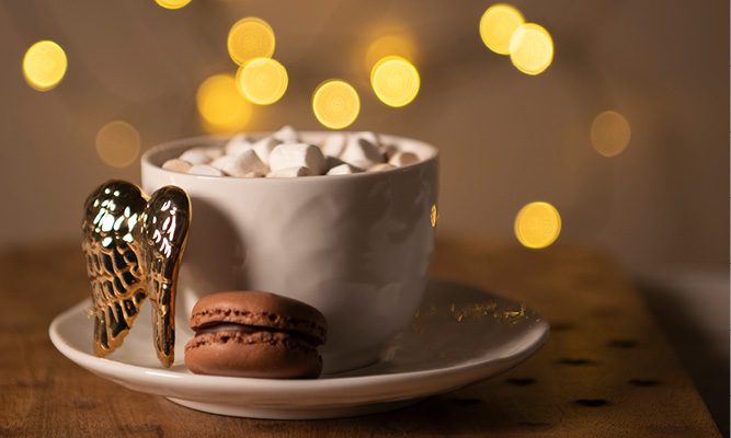 A cup and saucer filled with marshmallows and a brown macaron, with gold-colored wings as the handle