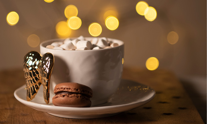 Una tazza e piattino riempiti di marshmallow e un macaron marrone, con ali dorate come manico