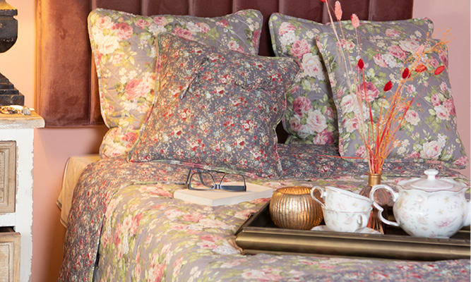 Een opgemaakt bed met romantische kussens en bedsprei en op het bed ligt een goudkleurige dienblad met theekopjes en theepot