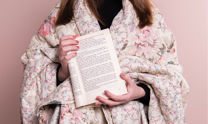 A person wrapped in a bedspread with a book in their hands