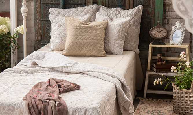 Une chambre à coucher shabby chic avec un lit fait avec des oreillers et une couvre-lit. À côté se trouve une table de chevet de style campagnard avec une horloge de table et un cadre photo