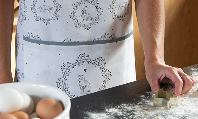 A woman is wearing a white apron with a gray cat motif, including a front pocket with gray trim. The motif depicts a sitting cat surrounded by a circle of branches. 