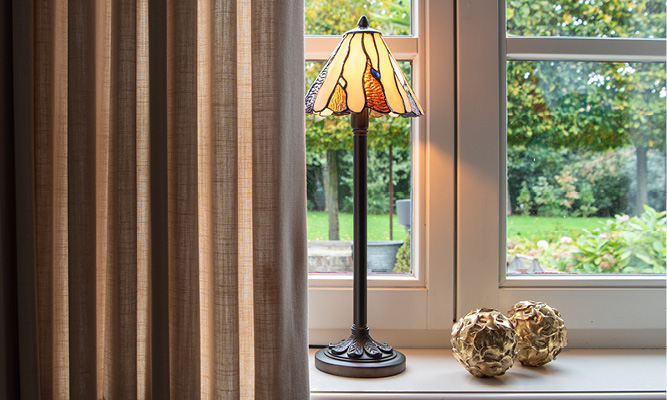 Een moderne vensterbank met een beige gordijn en op de vensterbank staat een antieke Tiffany tafellamp met twee goudkleurige decoratie bollen