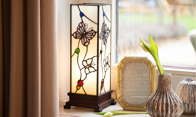 A Tiffany table lamp with butterflies on a windowsill with a gold photo frame, vases, and white tulips