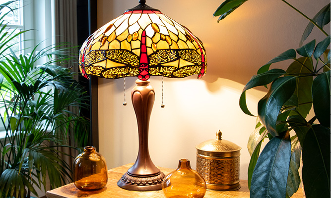 Een klassieke Tiffany tafellamp met een gele lampenkap met rode libelles en op het dressoir staan bruine glazen vaasjes en een zilverkleurig doosje