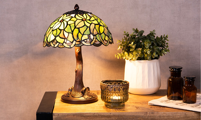 Une lampe de table Tiffany avec un motif de fleurs vertes, à côté se trouvent un porte-bougie en verre, un pot de fleurs blanc et deux bouteilles brunes
