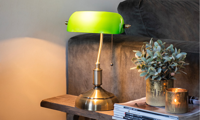 Une lampe de banquier avec un abat-jour vert sur un banc en bois avec des pots de fleurs et des magazines