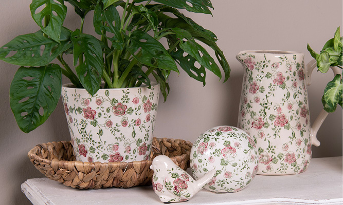 Een verzameling romantische woonaccessoires, waaronder een bloempot, schenkkan, vogel beeldje en decoratie bol met een roze bloemen motief