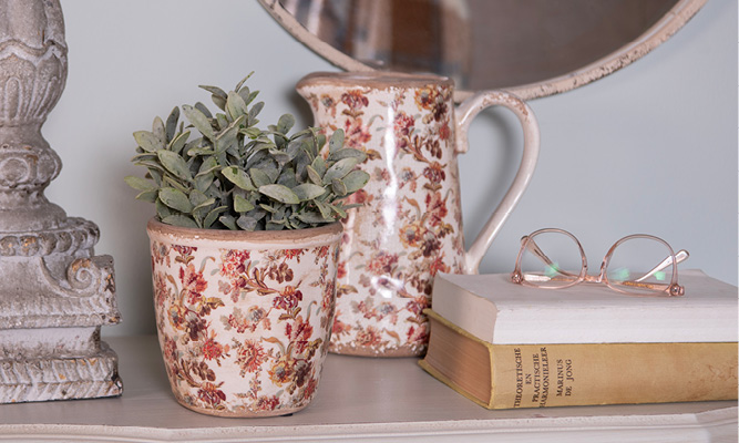 Een ouderwetse bloempot met een antiek bloemenpatroon, erachter staat een decoratieve schenkkan en ernaast liggen twee oude boeken