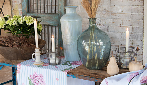 Un style d'intérieur champêtre avec un grand vase bleu avec des fleurs séchées brunes, une nappe d'hortensias, un vase métallique bleu clair et des bougeoirs dans des paniers métalliques