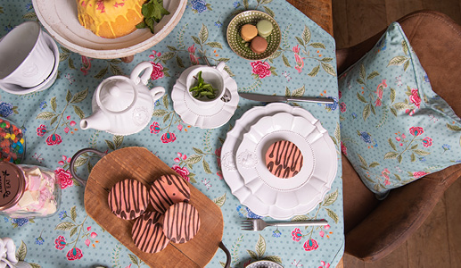 A cheerfully set table for a high tea party with a blue tablecloth featuring pink flowers and white romantic tableware and pink pastries