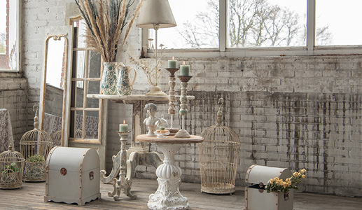Uno stile shabby chic con diversi oggetti tra cui tavolini, candele, gabbie per uccelli, casse, brocche e fiori secchi