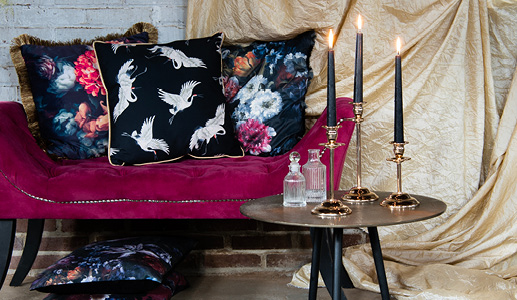 Ein antikes purpurfarbenes Sofa mit drei dramatischen Zierkissen auf dunklem Hintergrund und ein silberner Beistelltisch mit drei goldenen Kerzenständern und zwei Glasvasen