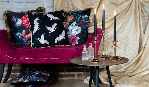 An antique purple sofa with three dramatic decorative cushions against a dark background and a silver side table adorned with three gold candelabras and two glass vases