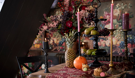 Une table richement dressée, garnie de fruits et de légumes, et une étagère noire avec des chandeliers noirs et dorés