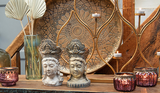 Two Buddha figurines with a large, colorful tray and a candle holder, alongside a green vase filled with dried flowers and two red glass tealight holders