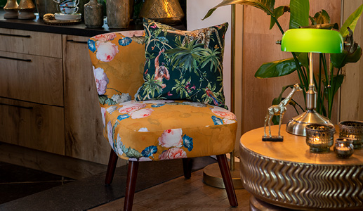 Un fauteuil jaune avec un coussin à motif jungle et une table d'appoint dorée avec une lampe de banquier verte Tiffany