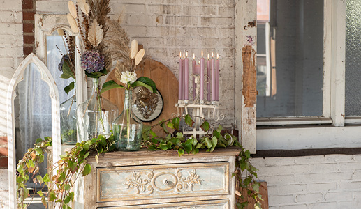 Eine brokante Kommode mit Kunstpflanzen, einem weißen Kerzenleuchter mit lila Abendkerzen und zwei großen Glasvasen mit einem Trockenblumenstrauß