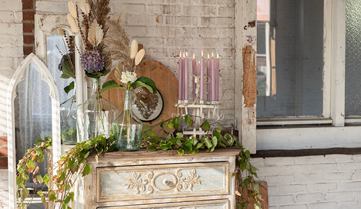 A brocante chest of drawers with artificial plants, a white candlestick with purple dinner candles, and two large glass vases with a dried flower bouquet