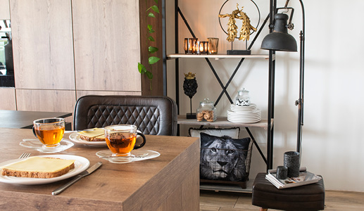 An industrial kitchen with two leather stools, glass tea cups, a large industrial floor lamp, and a shelving unit with cushions, tableware, figurines, and tea light holders