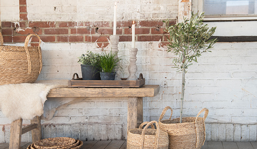 A weathered brick wall with a wooden bench adorned with a sheepskin, wooden tray, two anthracite flower pots, and two gray concrete candleholders, along with two wicker baskets with an olive tree