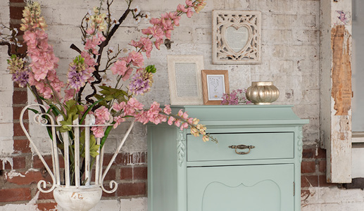 Une armoire de rangement vert menthe avec deux cadres photo et un porte-bougie, et un cadre photo accroché au mur, avec un vase en fer blanc avec des fleurs en faux rose à côté