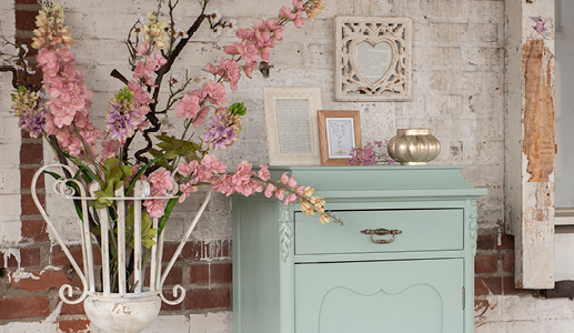 Een mintgroene opbergkast met twee fotolijstjes en een theelichthouder en een fotolijst aan de muur, aan de zijkant staat een witte ijzeren vaas met roze kunstbloemen