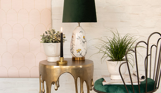 Een gouden bijzettafel met een gouden kandelaar en een witte bloempot met een gezicht en een modern tropische tafellamp met een zwarte lampenkap, vooraan staat nog een moderne bloempot met een minimalistisch gezicht