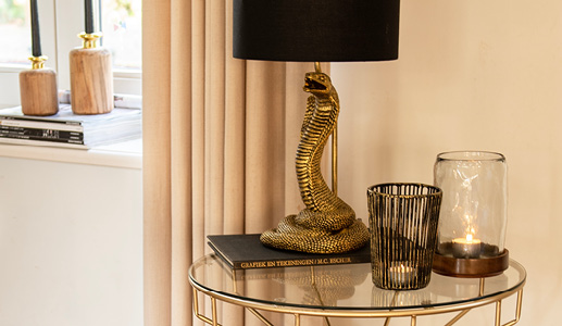 A gold side table with a glass top, featuring a gold cobra table lamp, a metal tealight holder, and a glass tea light holder, with a beige curtain in the background