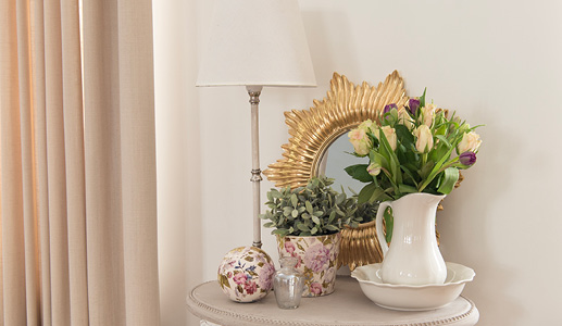 Un mur beige avec des rideaux beiges et une table d'appoint garnie d'un miroir mural doré, d'une lampe de table blanche, d'un pichet blanc, d'un pot de fleurs à motif floral et d'une sphère décorative