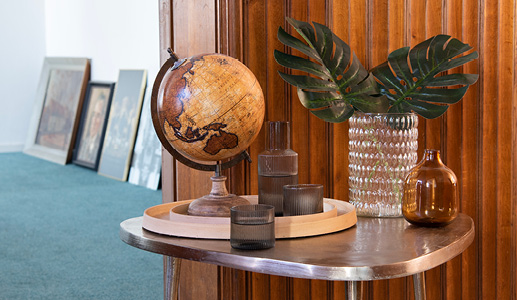 A silver side table with an antique brown world globe, a gray glass pouring jug, and drinking glass, along with a glass vase filled with artificial branches and a beige vase